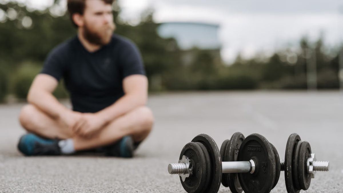 Young male with beard resting after workout on asphalt road with heavy dumbbells on gloomy day in selective focus